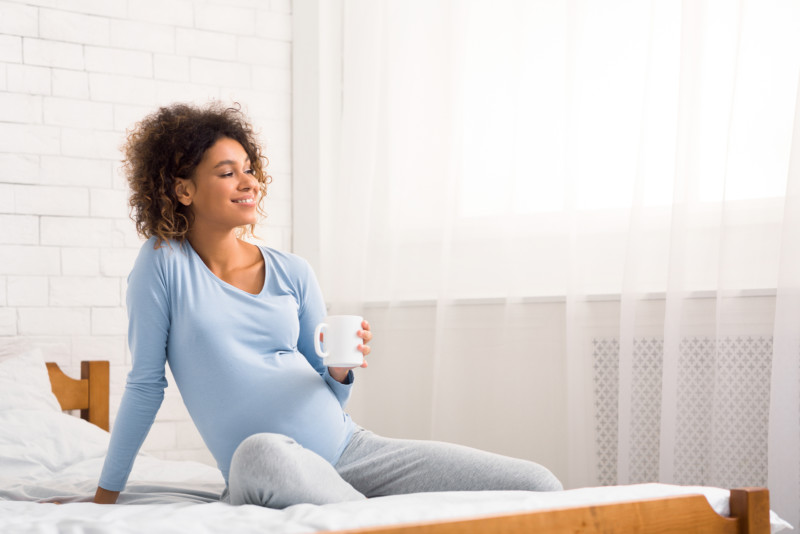 Schwangere Frau sitzt mit einer Tasse Himbeerblaettertee auf dem Bett